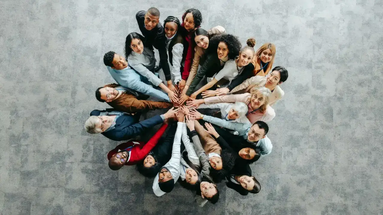 Grupa ludzi w kręgu, dłonie złożone na środku, symbolizująca współpracę przy wypełnianiu kwestionariusza osobowego dla osoby ubiegającej się o zatrudnienie.