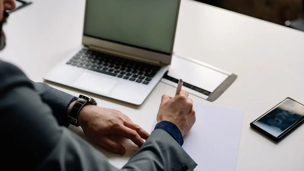 Mężczyzna w garniturze podpisuje dokumenty, co może oznaczać zamknięcie działalności gospodarczej. Obok leży laptop i telefon.