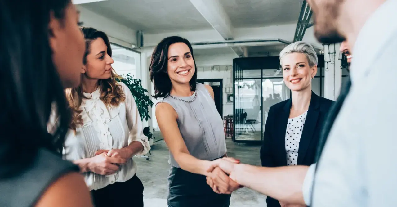 Uśmiechnięte kobiety w biurze, jedna z nich ściska dłoń partnerowi, symbolizując udaną współpracę w spółce partnerskiej.
