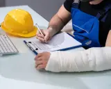 Pracownik z ręką w gipsie wypełnia dokumenty. Może to być formularz do ZUS odszkodowania, gdzie cennik i tabela pomogą w ustaleniu kwoty.