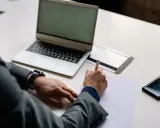 Mężczyzna w garniturze podpisuje dokumenty, co może oznaczać zamknięcie działalności gospodarczej. Obok leży laptop i telefon.