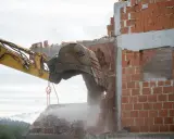 Koparka niszczy ceglaną ścianę. Rozbiórka postępuje, a gruz spada w dół.