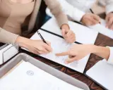 Dwie osoby podpisują dokumenty, jedna wskazuje palcem miejsce do podpisu. Może to dotyczyć formalności związanych z podatkiem od spadku po rodzicach.