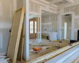 Wnętrze w trakcie przebudowy. Widać nowe drzwi, materiały budowlane, narzędzia i przygotowane do montażu listwy przypodłogowe.
