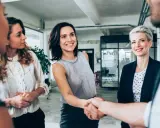 Uśmiechnięte kobiety w biurze, jedna z nich ściska dłoń partnerowi, symbolizując udaną współpracę w spółce partnerskiej.