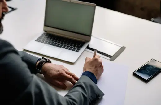 Mężczyzna w garniturze podpisuje dokumenty, co może oznaczać zamknięcie działalności gospodarczej. Obok leży laptop i telefon.