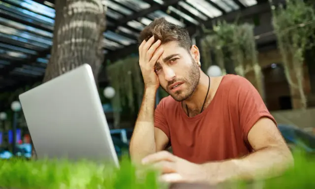Mężczyzna z ręką na czole, sfrustrowany pracą przy laptopie. Czyżby padł ofiarą pomówienia w internecie?