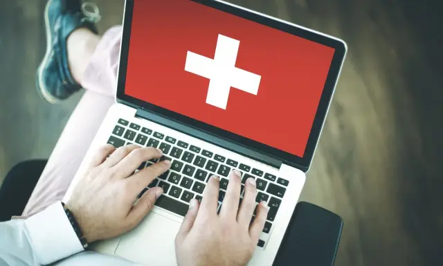 Osoba pisząca na laptopie z flagą Szwajcarii na ekranie.