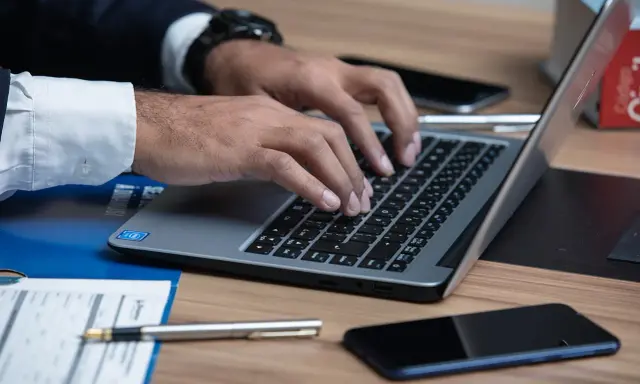 Prawnik pisze na laptopie, szukając informacji, jak napisać apelację od wyroku karnego wzór. Na biurku leżą dokumenty i telefon.