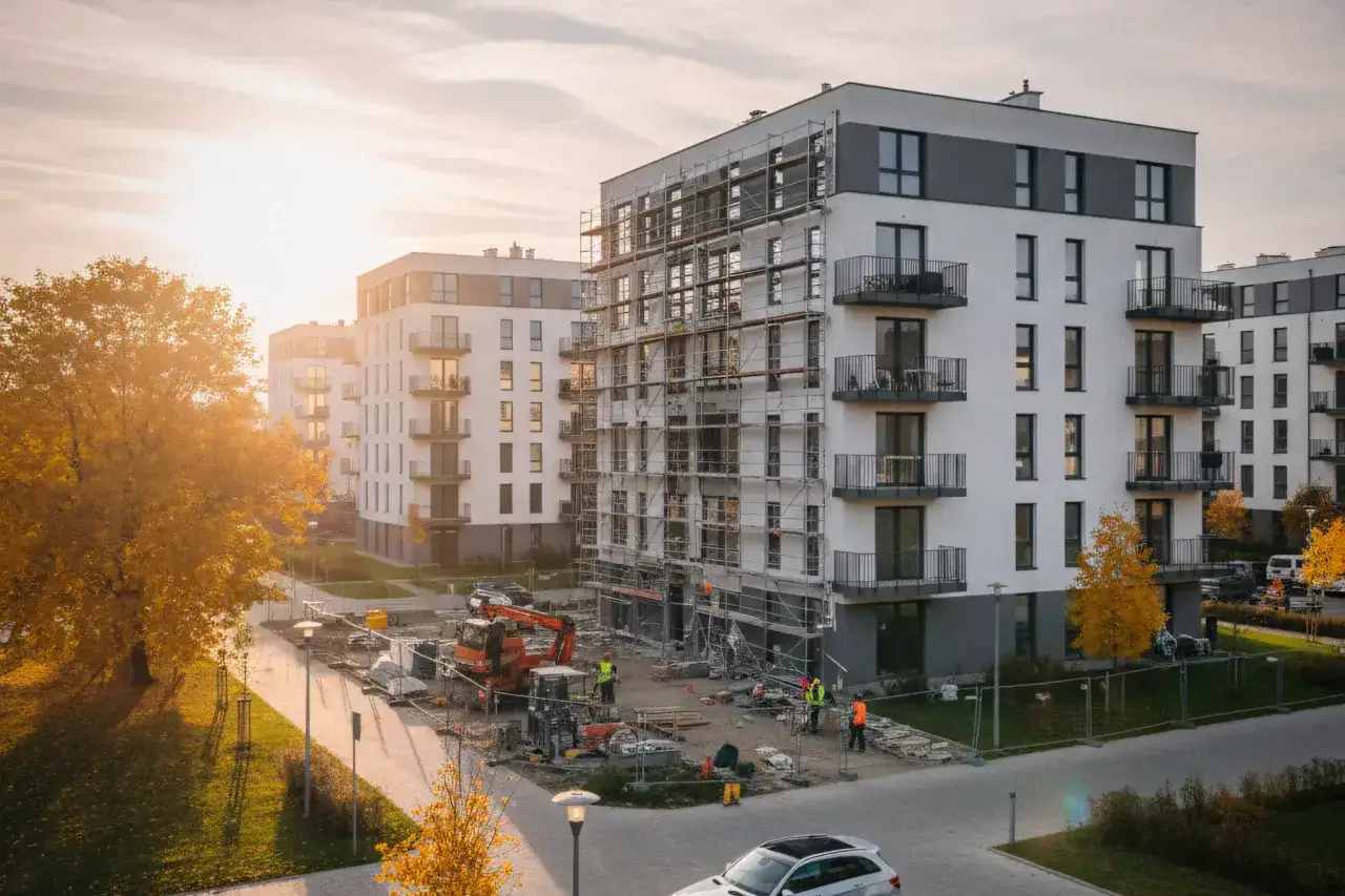 Budowa osiedla mieszkaniowego. Widać rusztowania na budynkach, koparkę i robotników. Jesienne drzewa dodają uroku. To miejsce, gdzie powstaje przyszła umowa deweloperska.