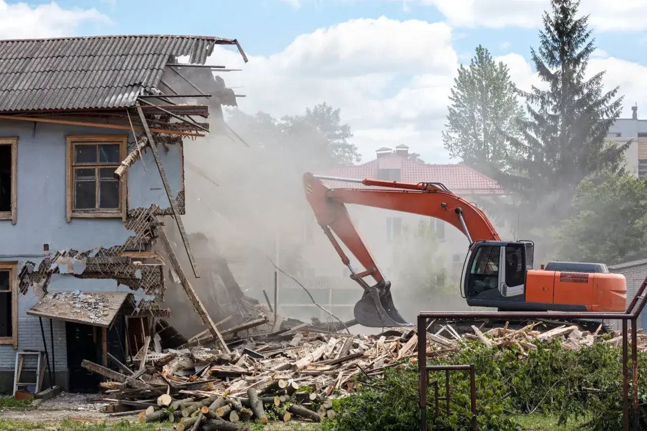 Koparka pracuje przy rozbiórce starego budynku, unosząc tumany kurzu.